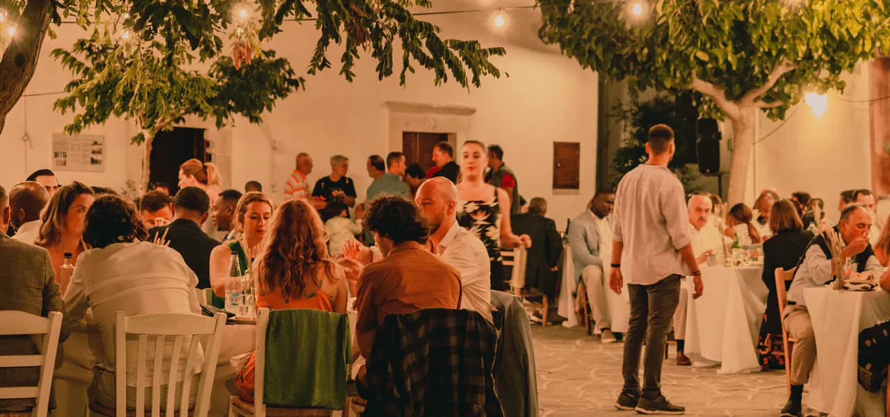Guests dining at tables in a lantern-lit courtyard at dusk, with trees overhead and neighbours chatting between the tables