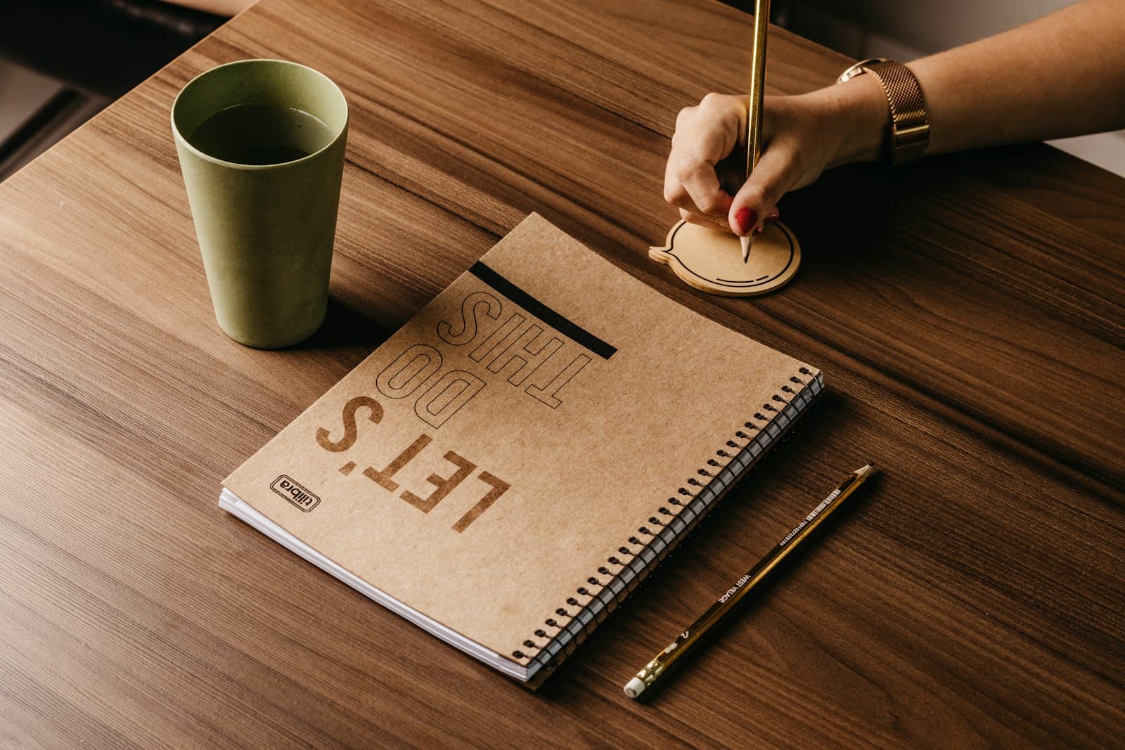 A person writes notes with a pen at a wooden cafe table next to a notebook and a cup of coffee.