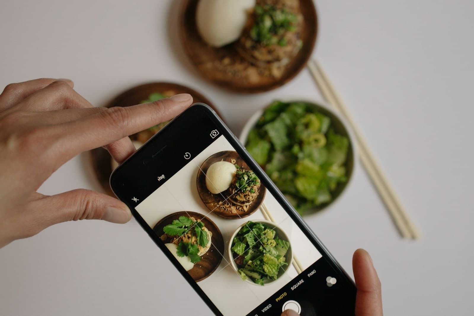 A hand frames a phone photo of bowls of food on a light table, seen from above.