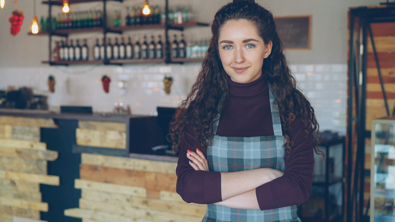 A smiling cafe owner in a plaid apron stands with her arms crossed in front of the counter of her coffee shop.