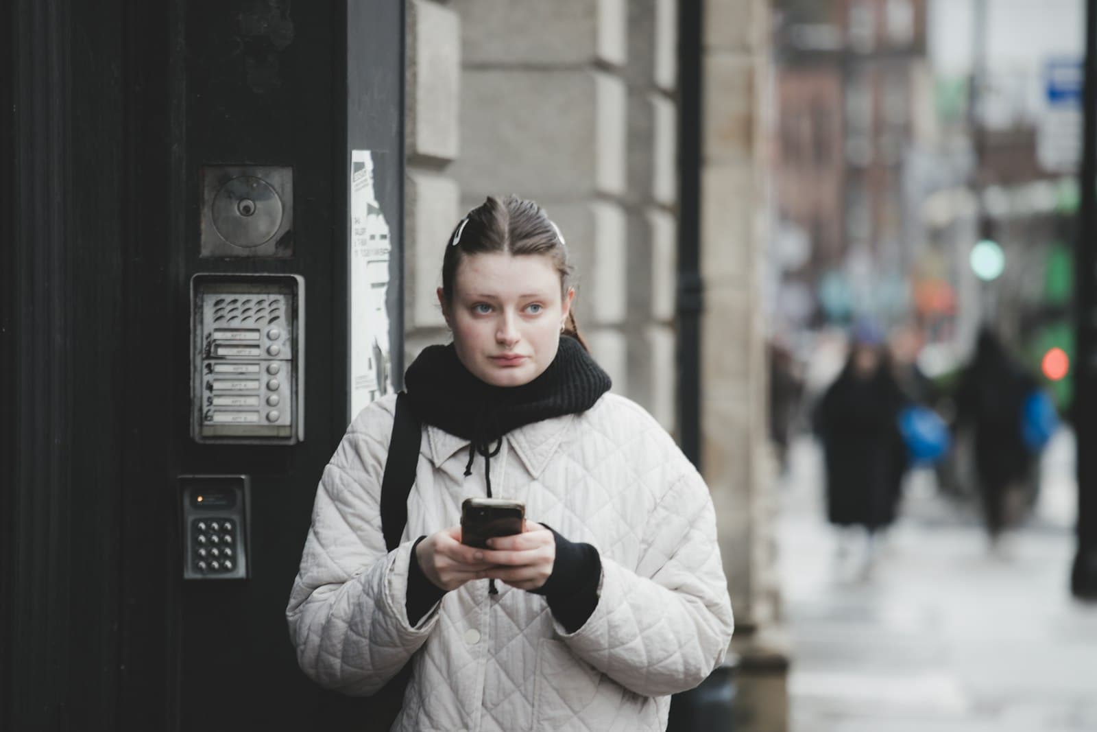 A young woman in a winter coat looks up from her phone on a busy city street.