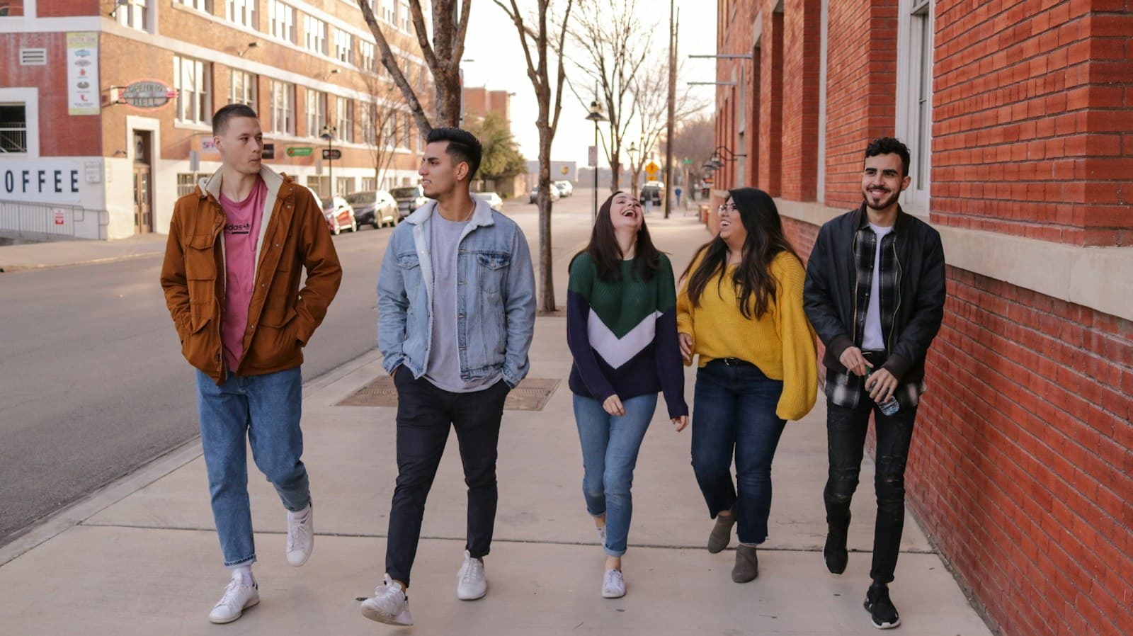Five friends walk and laugh together along a brick-lined city sidewalk.