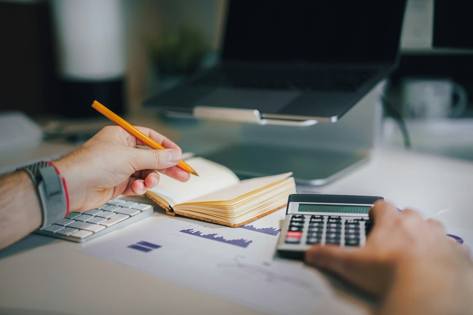 Hands working through figures with a pencil, an open notebook and a calculator on a desk beside a laptop.